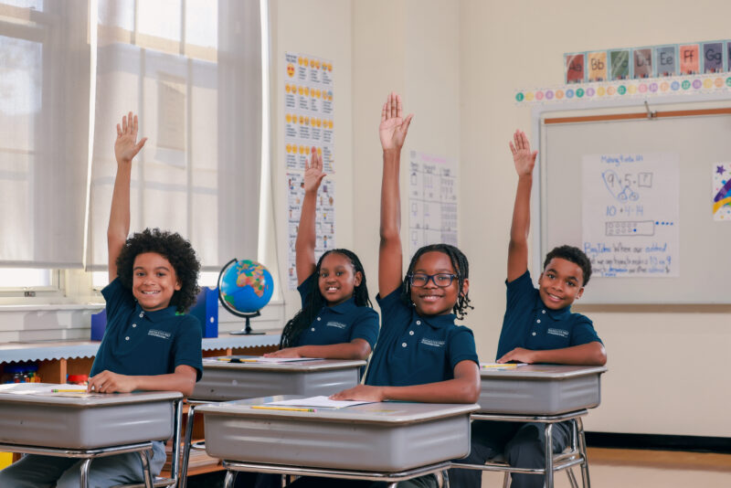 Students sitting at desks raising hands and smiling